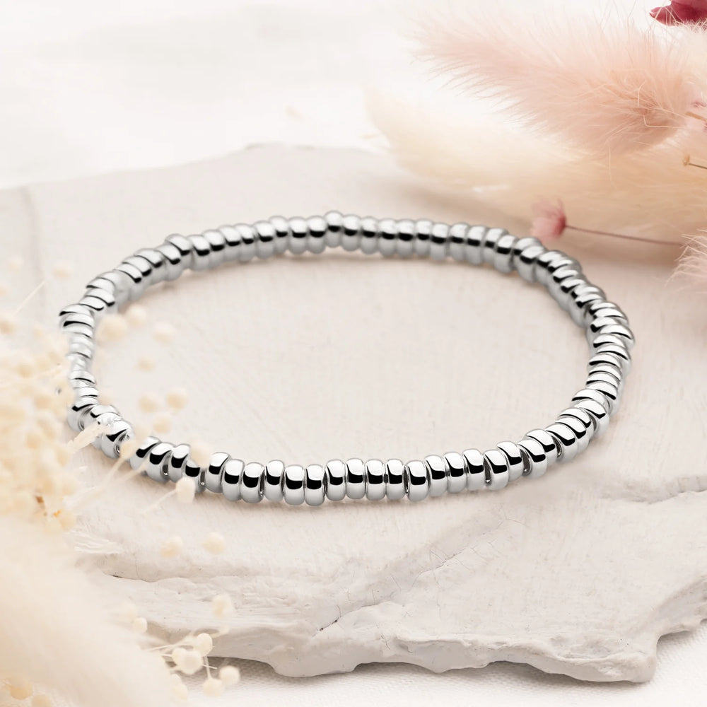 A silver bracelet with a pebble-like texture, displayed on a stone surface, with a neoprene band that allows for adjustable expansion.