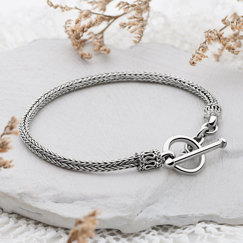 A sterling silver bracelet with a classic Balinese weave design and a decorative T-Bar closure, displayed on a white surface with dried flowers in the background.