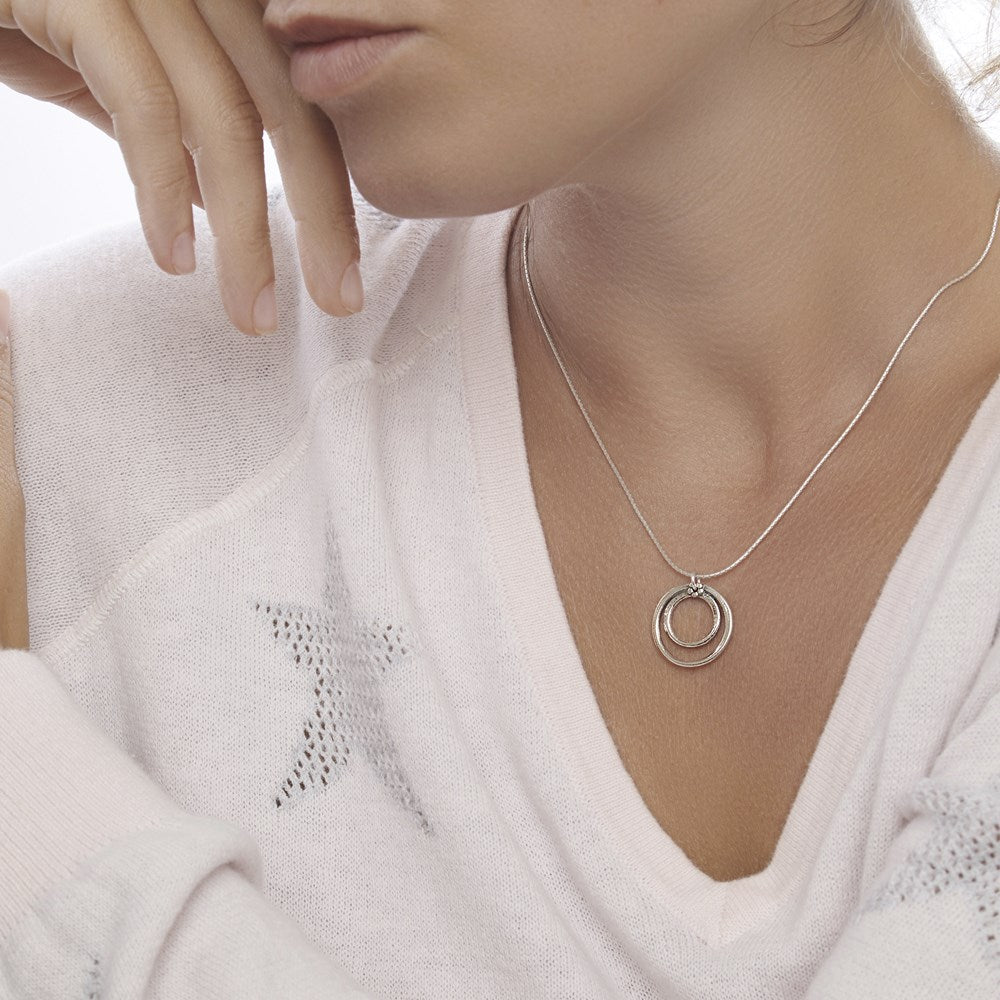 A woman model wearing a silver necklace with two nested hoops pendant attached to a chain, displayed against a wooden background.