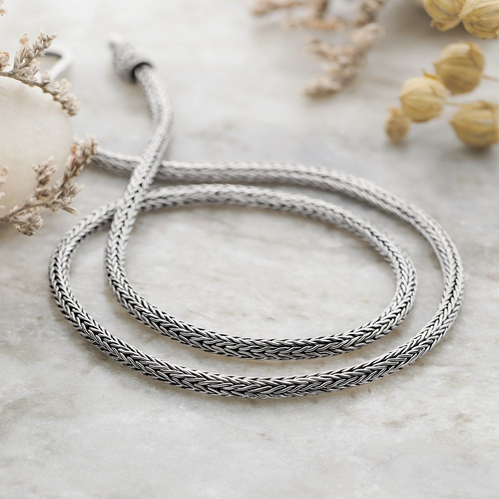 A close-up image of a handmade Balinese silver chain necklace with a classic weave pattern, displayed on a marble surface.