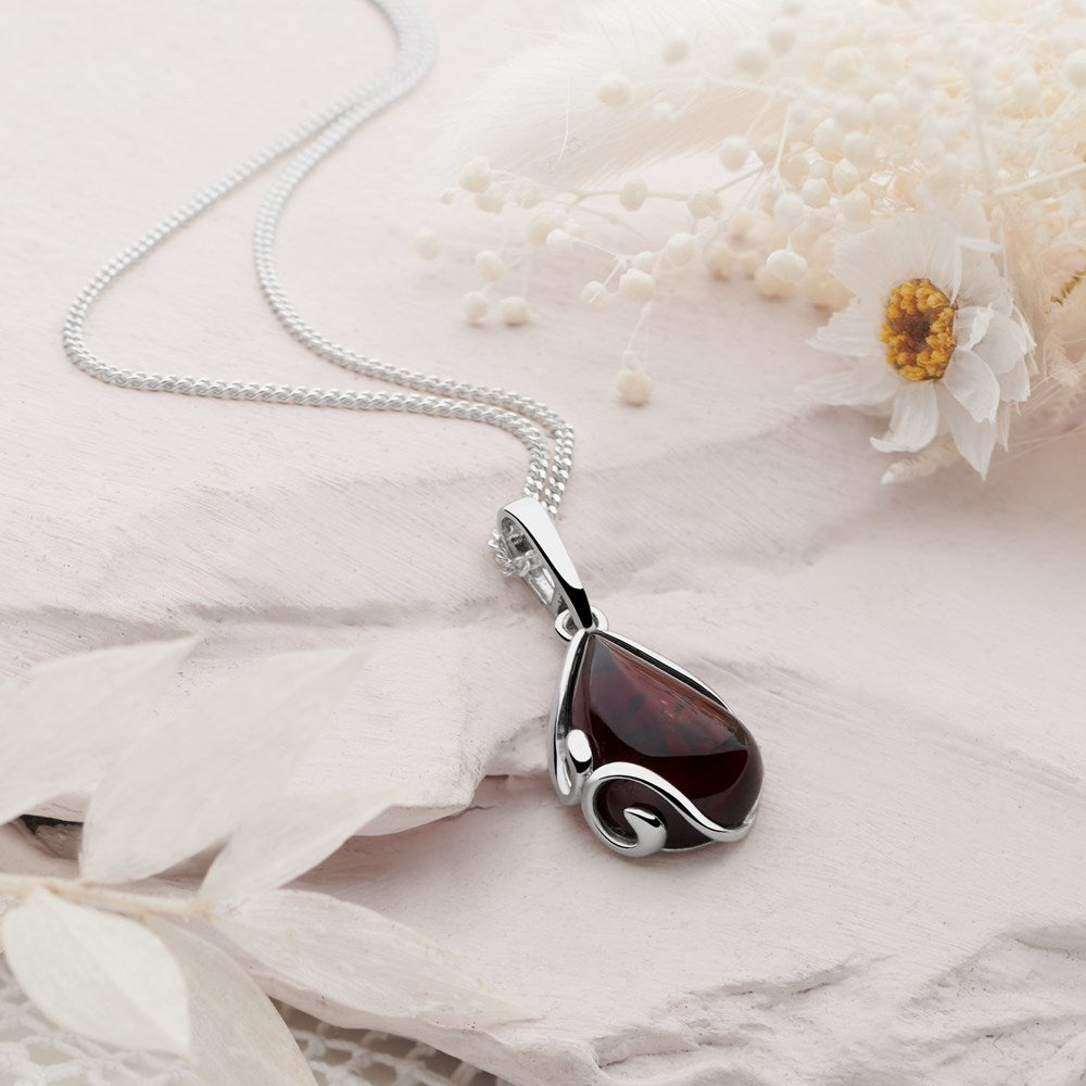 A teardrop-shaped amber pendant set in silver, accompanied by a 40cm curb chain, displayed on a white textured surface with dried flowers.