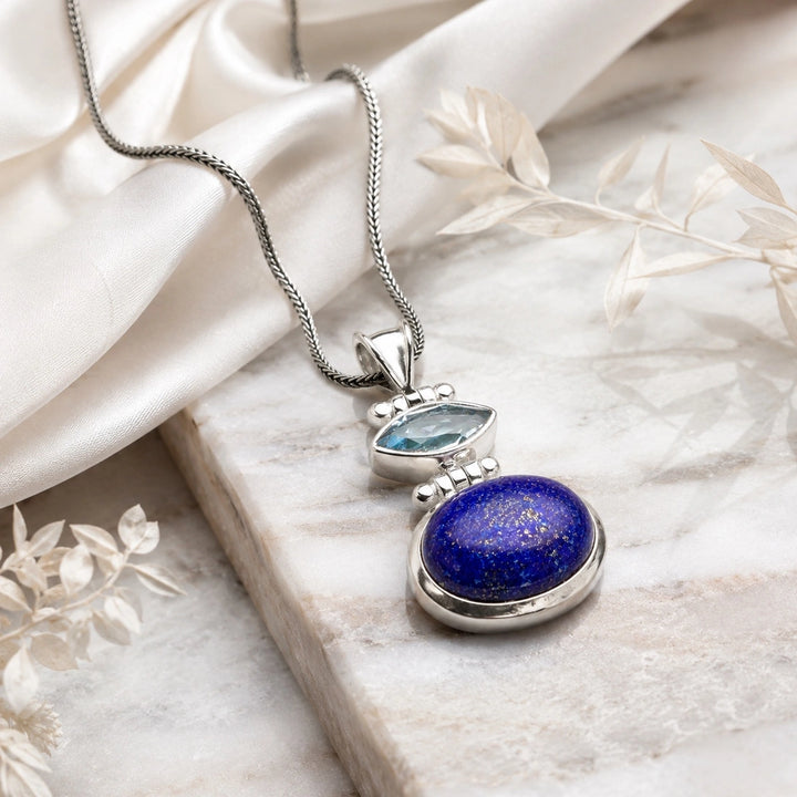 An intricate silver pendant featuring a marquise-cut light blue gemstone set above a larger oval deep blue lapis lazuli stone, suspended from a textured silver chain.