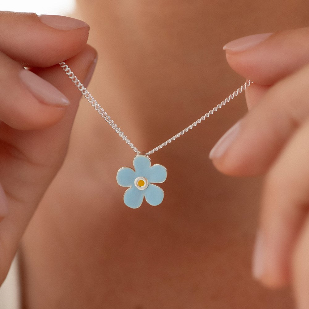 A close up image of blue petal-shaped pendant with a white and yellow center, attached to a silver curb chain held by woman. 
