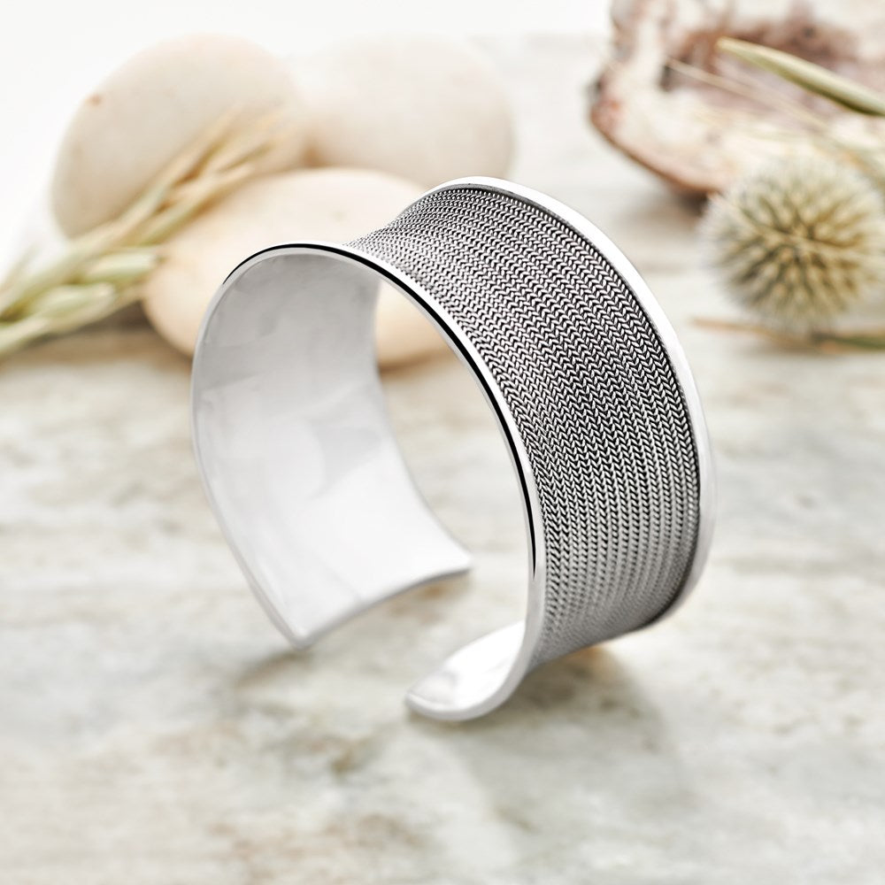A silver bangle bracelet with intricate filigree wire work, displayed against a backdrop with neutral colors.
