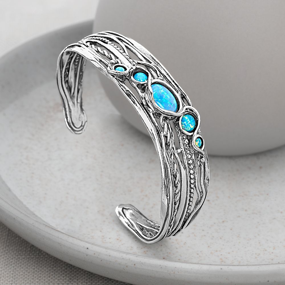 A silver bangle bracelet with blue opal stones set in an openwork design, displayed on a white plate.