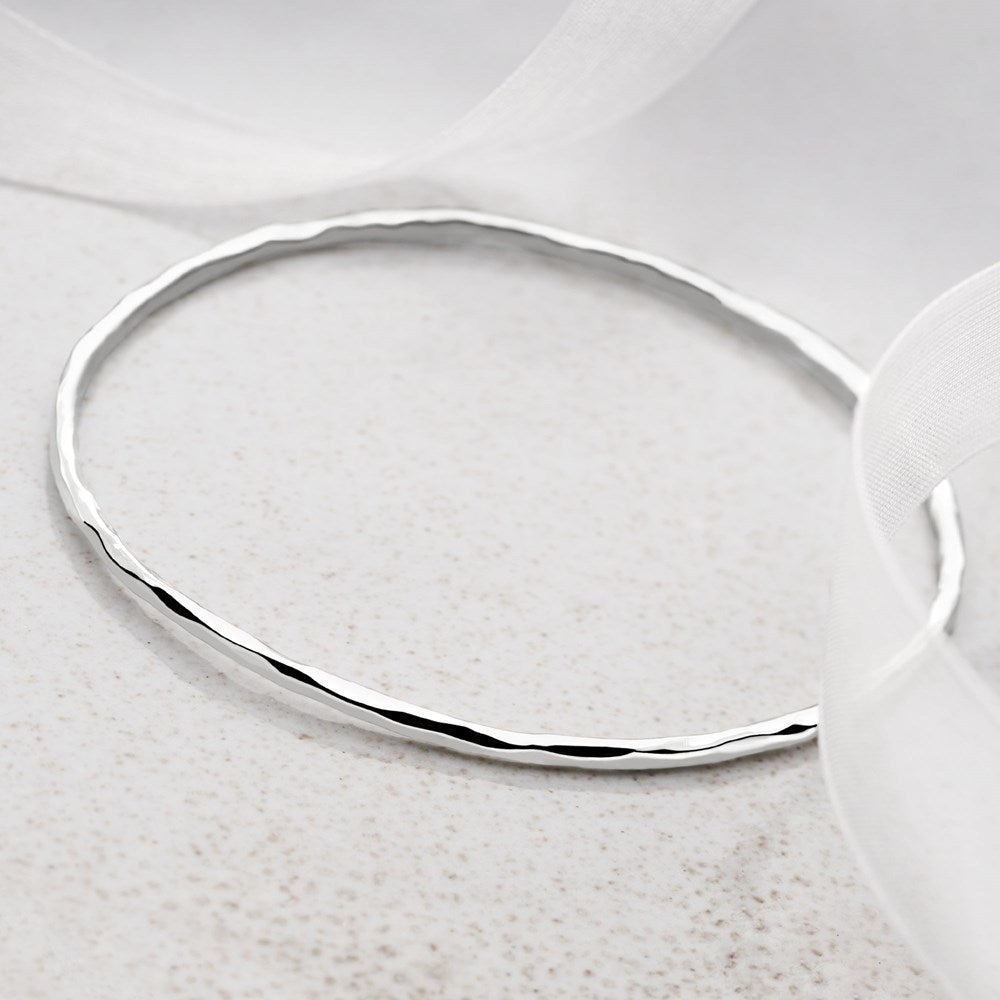 A silver bangle bracelet with a hammered finish, displayed on a white surface with a reflective shadow.