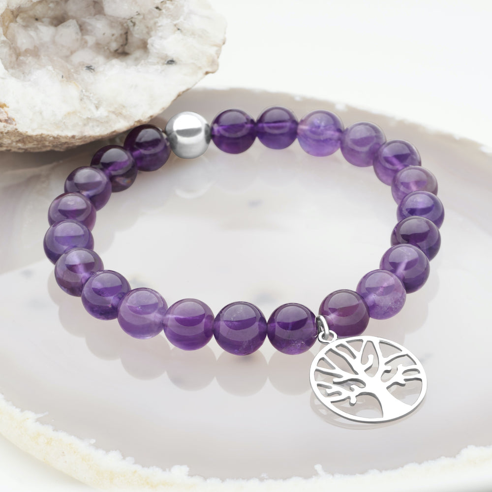 A bracelet made of purple amethyst beads with a silver tree of life charm, displayed on a white surface.