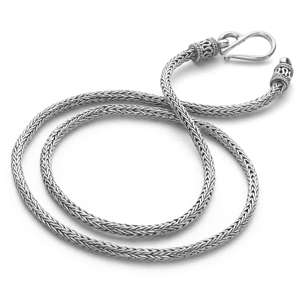 A silver-colored Balinese chain necklace laid out on a marble surface, displaying its intricate weave pattern.