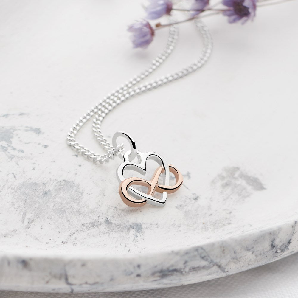 A sterling silver necklace with a rose gold-plated heart and infinity symbol pendant, displayed on a marble surface with a silver curb chain.
